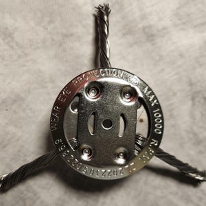 Puede incluir: Primer plano de un accesorio de cable de acero con una placa circular. La placa tiene el texto "WEAR EYE PROTECTION" y "MAX 10000 R.P.M.". El accesorio está unido a tres cables de acero.