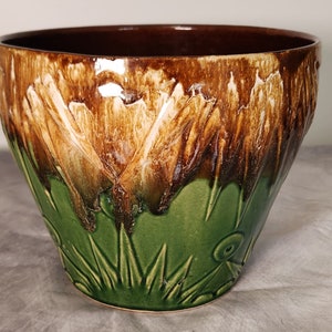 Może przedstawiać: Gliniana doniczka w brązowo-zielonym szkliwie z kwiatowym wzorem. Doniczka ma szeroki rant i zaokrąglone dno.