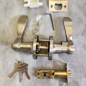 May include: A silver door handle set with a brushed nickel finish. The set includes a door handle, a deadbolt, a strike plate, a set of keys, and screws. The door handle has a lever design and the deadbolt has a round knob. The set is packaged in a clear plastic bag.