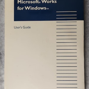 Puede incluir: Una Guía del usuario de Microsoft Works para Windows sellada. La portada es blanquecina con una sección rectangular azul oscuro en la parte superior. El texto "Microsoft Works para Windows" es blanco, y "Guía del usuario" está debajo.