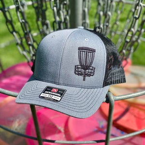 Könnte beinhalten: Graue und schwarze Trucker-Kappe mit einem schwarzen, gestickten Disc-Golf-Korb auf der Vorderseite. Die Kappe hat ein schwarzes Netzrücken und ein schwarzes Etikett mit dem Text "Richardson 112" auf der Vorderseite.