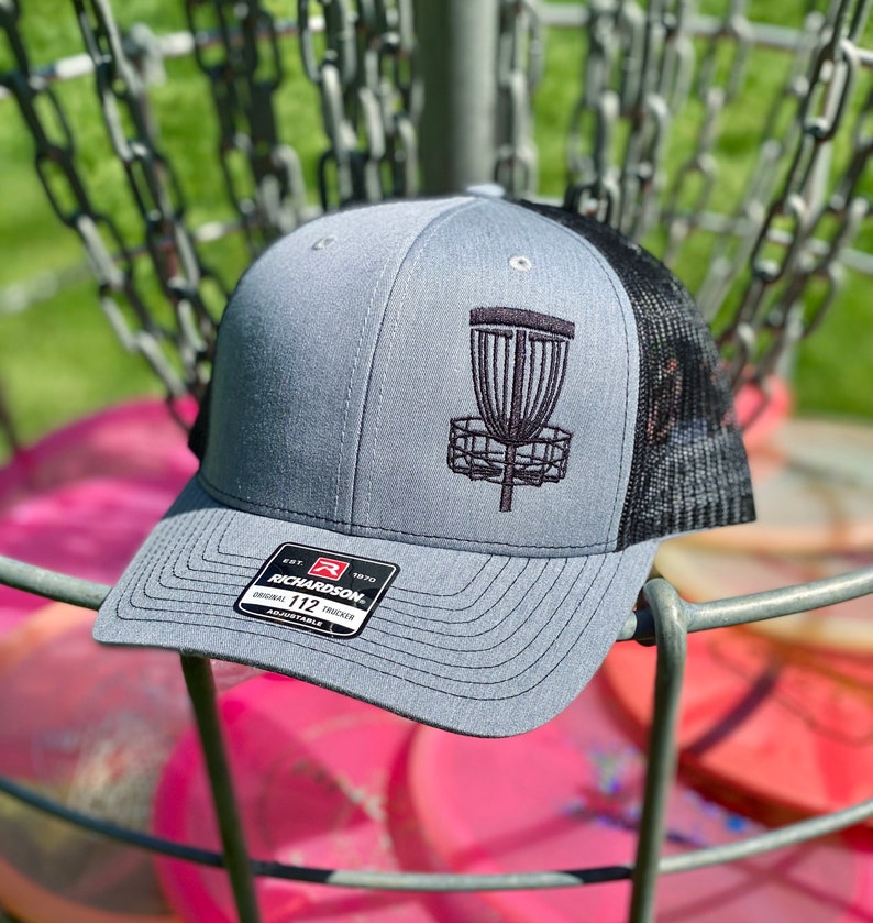 May include: Gray and black trucker hat with a black embroidered disc golf basket on the front. The hat has a black mesh back and a black label with the text "Richardson 112" on the front.