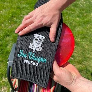 Puede incluir: Toalla de disco de golf negra con un diseño de cesta de disco de golf blanco y el texto "Jon Vaughn #96580" en bordado turquesa.