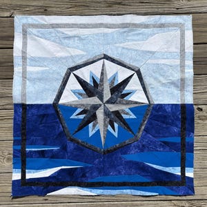 May include: A square quilt featuring a nautical compass design. The quilt has a blue and white color scheme, with a compass rose in the center. The background depicts a sea and sky scene, with a dark blue border.