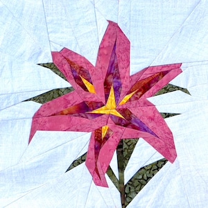 May include: A close-up of a fabric flower quilt block. The flower has pink petals with purple and burgundy accents, a yellow center, and green leaves. The background is white with light blue lines.