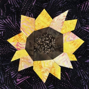 May include: A close-up of a quilt block featuring a sunflower design. The sunflower is made of yellow, orange, and pink fabric, and the center is a dark brown fabric with a swirling pattern. The background is a dark purple and black fabric with a swirling pattern.