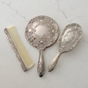 May include: A vintage vanity set featuring a round hand mirror, a hairbrush, and a comb. The set is silver-toned with ornate floral detailing. The comb has a cream-colored plastic comb. The mirror and brush have handles.