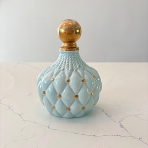 Puede incluir: Frasco de perfume de vidrio azul claro acolchado con un tapón esférico dorado. La botella tiene forma redondeada con detalles de puntos dorados. El tapón y el cuello de la botella también son dorados.