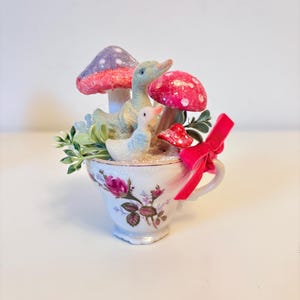 Op de afbeelding: Een wit theekopje met roze rozen en groene bladeren is gevuld met een kleine witte vogel, een groene eend en drie roze en witte paddenstoelen.