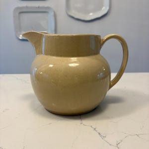 Puede incluir: Una jarra de cerámica beige con cuerpo redondeado, pico vertedor y asa curvada. La jarra tiene un acabado moteado y está sobre una superficie blanca. Platos decorativos blancos cuelgan en el fondo.