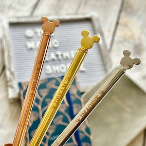 May include: Three gold-toned pens with Mickey Mouse head toppers. The pens are engraved with the words "Believe", "Dream", and "Imagine".