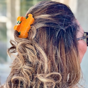 May include: An orange hair clip shaped like a bear is nestled in wavy, light brown hair. The clip has green accents and is positioned near the back of the head. The hair is styled with loose curls and highlights.