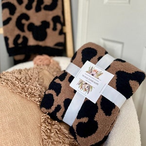 May include: A brown and black leopard print throw blanket with a white ribbon tied around it. The blanket is sitting on a white chair with a brown and tan fringed throw pillow. The label on the blanket reads "The Magic Feather Shoppe".