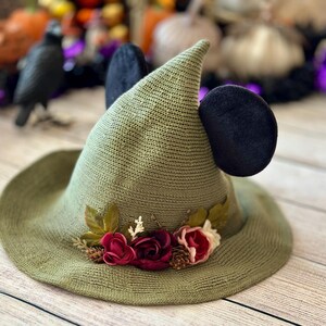 May include: A green witch hat with black mouse ears and a floral arrangement. The hat has a wide brim and a pointed crown. The floral arrangement includes burgundy and pink roses, small pinecones, and green leaves. The hat is made of a woven material.