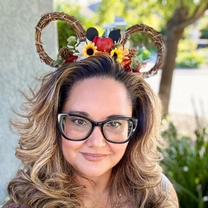 May include: A woman wearing a brown and tan headband with fall-themed decorations, including sunflowers, apples, and leaves. She is also wearing black glasses and a layered gold necklace.