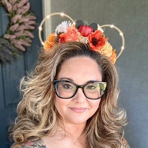 May include: A floral headband with orange, red, and white flowers, small black mouse ears, and a string of lights shaped like Mickey Mouse ears. The headband is worn on a person's head.