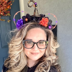 May include: A black witch hat headband with purple and orange flowers, black and gold accents, and a light-up wire halo. The headband is worn by a person with long blonde hair.