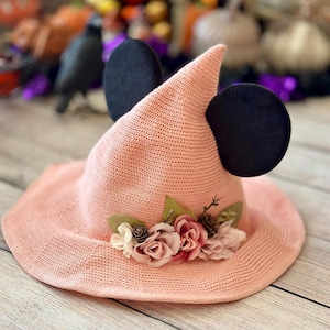 May include: A light pink witch hat with black mouse ears and a floral arrangement. The hat has a wide brim and a pointed crown. The flowers are pink and white, with green leaves. A fun accessory for Halloween or costume parties.