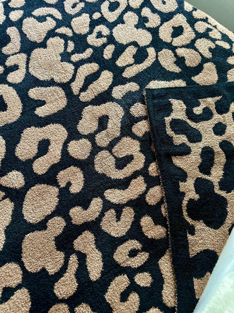 May include: A black and beige throw blanket with a leopard print pattern. The blanket is made of a soft, plush material.