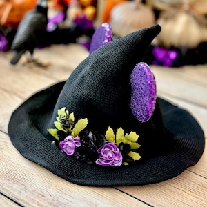 May include: Black witch hat with a wide brim, decorated with purple flowers, green leaves, and a small black spider. The hat has two purple sequined ear-shaped accents. A festive Halloween accessory.
