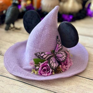 May include: A lavender witch hat with black mouse ears and a butterfly and floral arrangement. The hat has a wide brim and a pointed crown. The butterfly is purple and black, and the flowers are pink and purple.