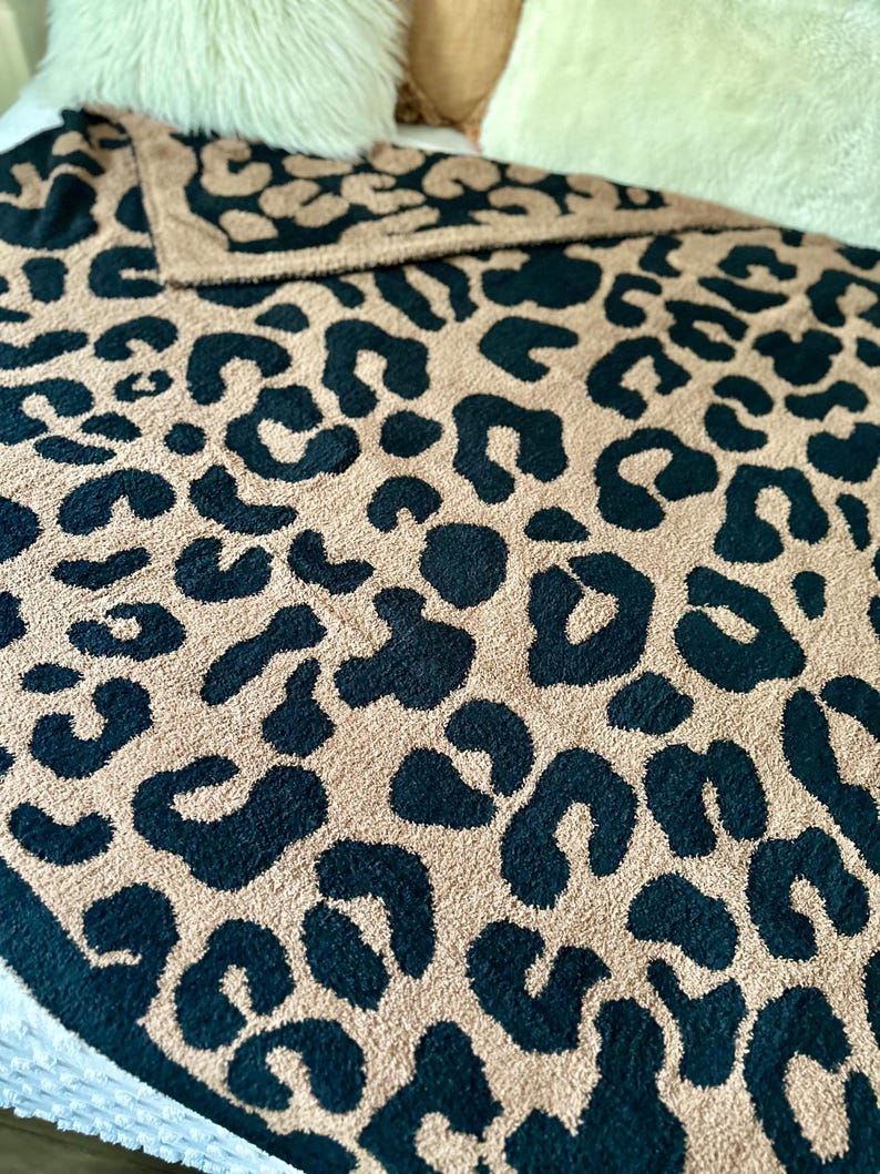 May include: A soft, plush throw blanket with a leopard print pattern in black and tan colors. The blanket is folded and displayed on a bed.