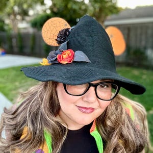 May include: A black witch hat with a wide brim, decorated with a red and orange rose, gray leaves, and black flowers. The hat is part of a Halloween costume. The hat is worn by a person wearing black glasses.