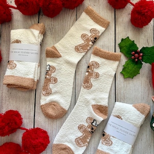 May include: Two pairs of white fuzzy socks with brown gingerbread man designs. The socks have a tan cuff and are packaged with a label that reads "The Magic Feather Shoppe".
