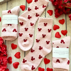 May include: Two pairs of pink fuzzy socks with red Mickey Mouse silhouettes. The socks are on a white wooden surface with red hearts scattered around them. The socks have a tag that reads "The Magic Feather Shoppe".