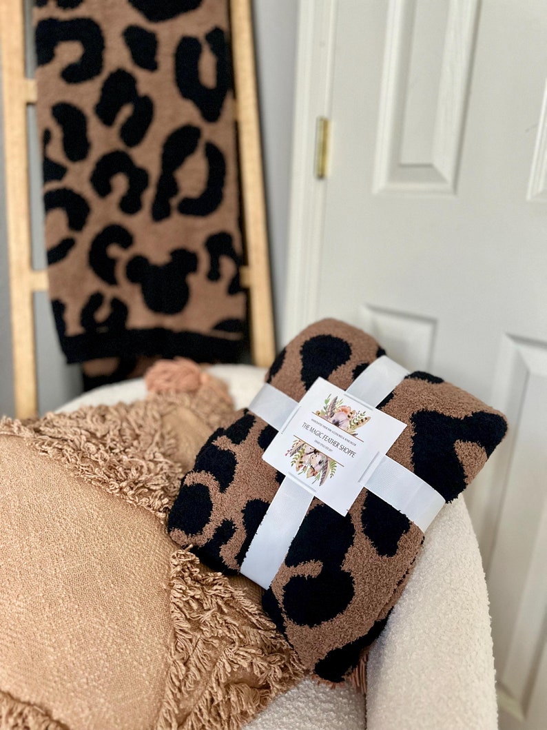 May include: A brown and black leopard print throw blanket with a white ribbon tied around it. The blanket is sitting on a white chair with a brown and tan fringed throw pillow. The label on the blanket reads "The Magic Feather Shoppe".