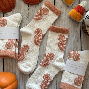 May include: Pair of white fuzzy socks with a brown Mickey Mouse design. The socks are on a wooden surface with fall decorations, including pumpkins and candy corn. The text "The Magic Feather Shoppe" is on a white tag.