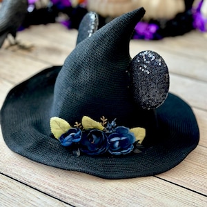 May include: A black witch hat with a wide brim, adorned with navy blue fabric flowers and light green leaves. The hat features sequined ears and is designed for Halloween or costume events.