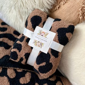 May include: A brown and black leopard print throw blanket with a white ribbon tied around it. The ribbon has a tag that says "The Magic Feather Shoppe".