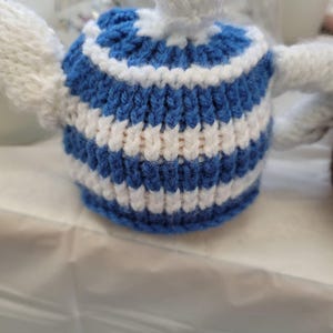 Può includere: Un copriteiera lavorato a mano a maglia con strisce blu e bianche. Il copriteiera ha una forma arrotondata con un piccolo pomello sulla parte superiore e due piccole maniglie. Progettato per adattarsi a una teiera standard e mantenere il tè caldo.