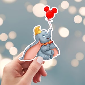 May include: A gray elephant with large pink ears holding a red balloon with Mickey Mouse ears. The elephant is wearing a yellow hat and a red and yellow striped collar.