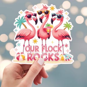 May include: A sticker featuring three pink flamingos wearing sunglasses, with the text "OUR FLOCK ROCKS." The flamingos are standing on a beach with palm trees and beach balls. The sticker has a white border.