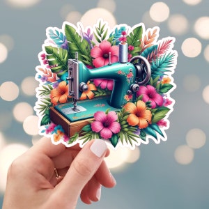May include: A teal sewing machine sticker surrounded by colorful tropical flowers and foliage. The sticker features a vintage-style sewing machine with floral accents, set against a backdrop of pink, orange, and green leaves and flowers. The sticker is held by a hand.