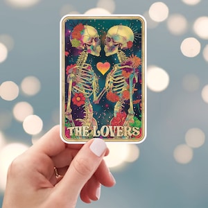 May include: A sticker featuring a tarot card design with two skeletons holding hands, surrounded by flowers and a starry background. The card reads "THE LOVERS" in a decorative font, with a gold border.