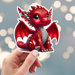 May include: A red dragon sticker with large, sparkling red gems covering its body. The dragon has large, expressive eyes, silver accents, and red wings. The sticker is held by a hand against a blurred background.