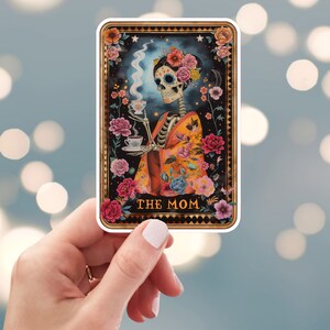 May include: A tarot card with a black background and gold trim. The card features a skeleton wearing a colorful kimono and holding a teacup. The card is titled "THE MOM".