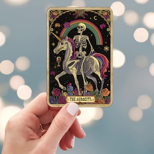 May include: A tarot card illustration featuring a skeleton riding a unicorn under a rainbow. The card has a black background with floral and celestial accents. The text "THE AUDACITY" is at the bottom.