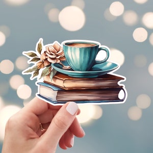 May include: A teal blue coffee cup and saucer sitting on top of a stack of three books. The books are brown, blue, and brown. A white flower with green leaves is behind the cup.