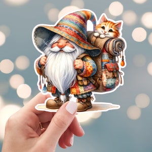 May include: A whimsical sticker featuring a smiling gnome with a long white beard, wearing a colorful hat and patchwork clothing. A ginger kitten peeks out from a backpack on the gnome's back. The sticker is held by a hand against a blurred background.