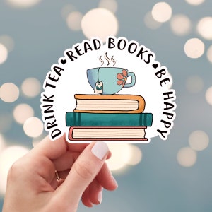 May include: A white sticker with the text "Drink Tea, Read Books, Be Happy" in black. A blue teacup with a flower design sits on top of a stack of three books.
