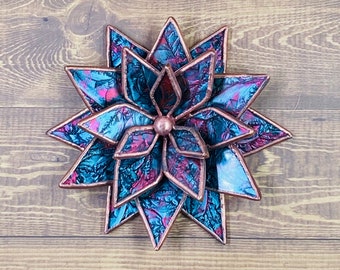 Puede incluir: Una flor de vidrio tintado de múltiples capas, tono cobre, con matices de azul verdoso y rosa. La flor tiene una forma de estrella estrellada con pétalos puntiagudos.