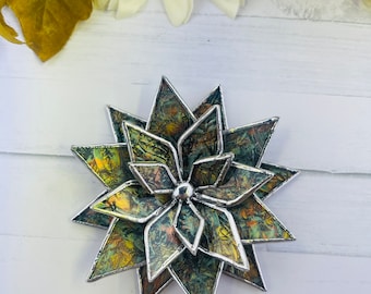 Puede incluir: Una flor de vidriera multicapa hecha a mano con un borde de metal plateado. El vidrio presenta una mezcla de colores verdes, amarillos y marrones. La flor mide aproximadamente 10 cm de diámetro, con un perno central plateado.