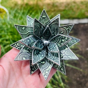 Puede incluir: Una flor de vidrio tintado verde y plateado con un diseño de estrella. La flor está hecha de múltiples capas de vidrio, cada una con un patrón diferente. La flor se sostiene en una mano.