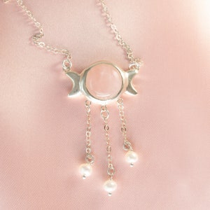Puede incluir: Un collar de plata con un colgante de piedra lunar rosa y tres abalorios de perlas que cuelgan de la parte inferior.