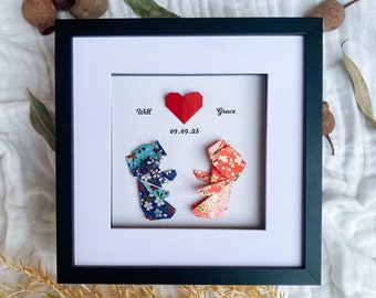 May include: Framed artwork featuring origami figures and a red paper heart. The black frame surrounds a white mat with the names "Will" and "Grace" and the date "09.09.23". The origami figures are in blue floral and red floral patterns.