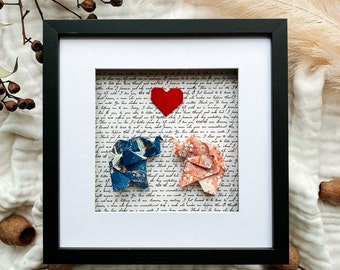 May include: Framed artwork featuring two origami elephants facing each other, a red heart, and a handwritten letter background. The elephants are blue and pink with floral patterns. The frame is black with a white mat.